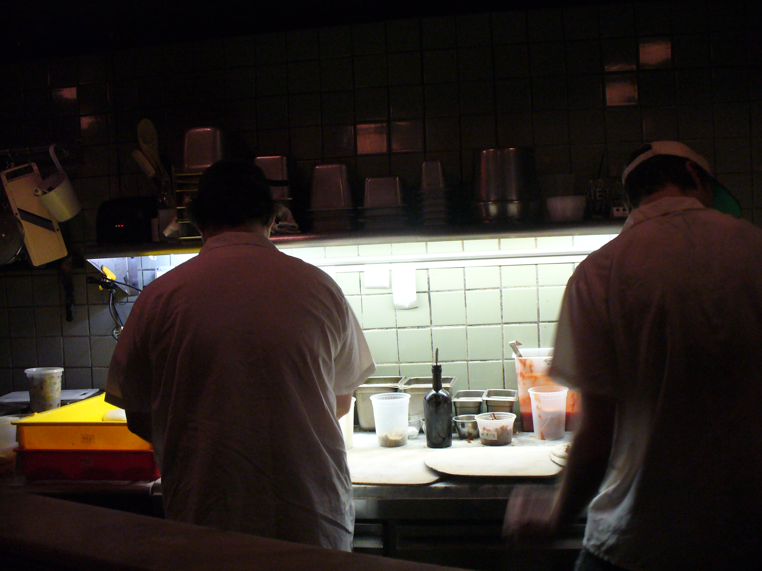 Two cooks at work in a low-lit kitchen, backs to the camera, prep counter glowing under a service light.